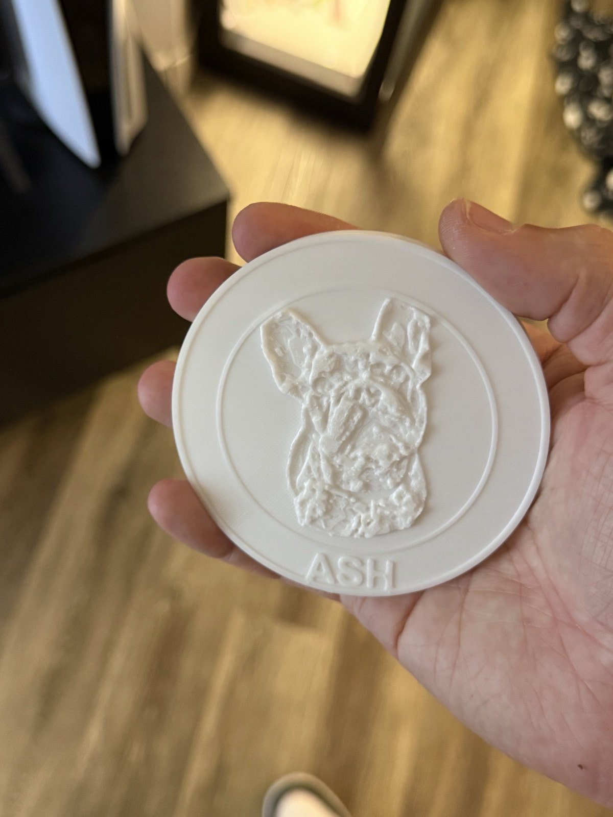 A white 3D-printed round coaster showing a relief portrait of a French bulldog and the name ASH along the bottom edge, held in a hand