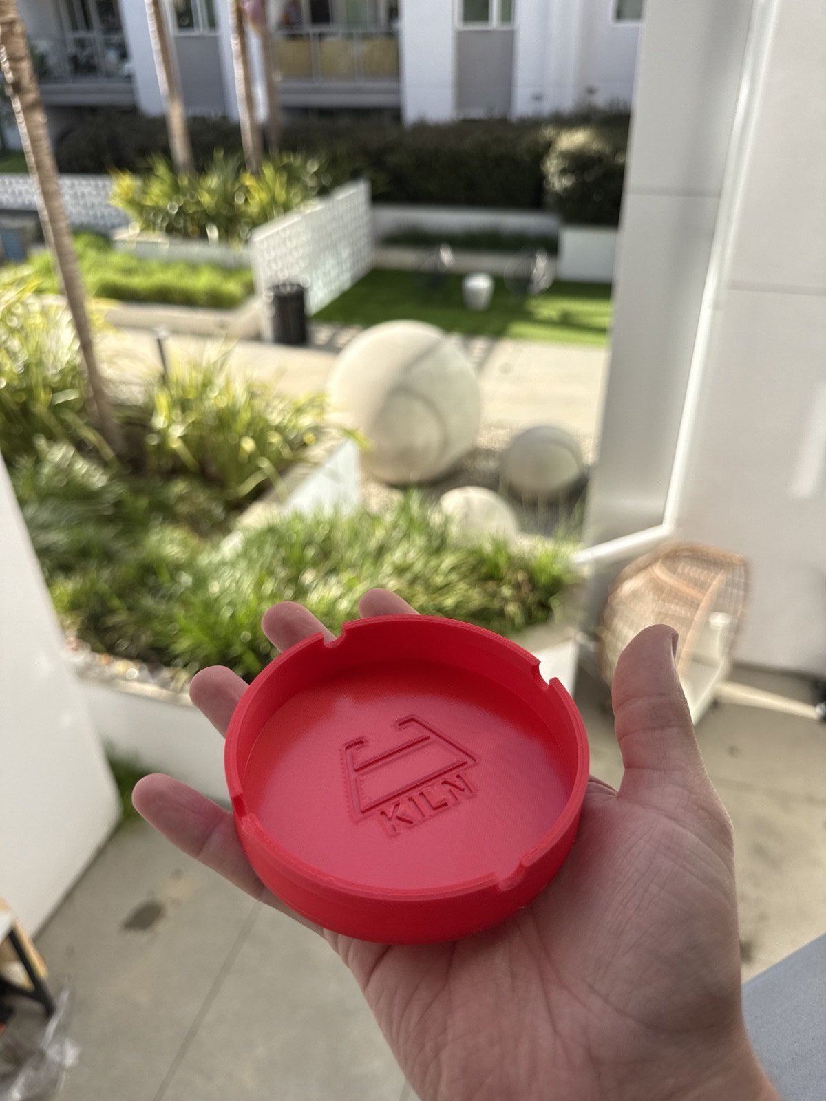 A red 3D-printed circular ashtray held in a hand, with the Kiln mark debossed into the inside